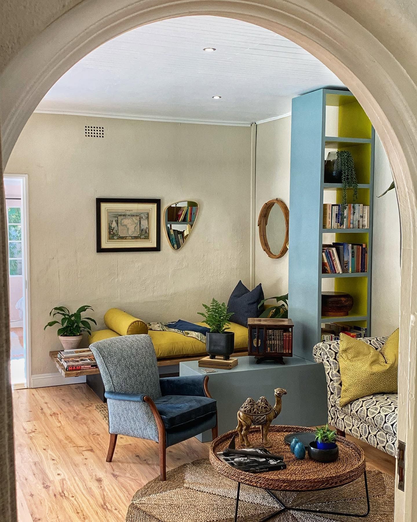A lounge seen through an arch: patterned chaise, painted shelves, rattan table